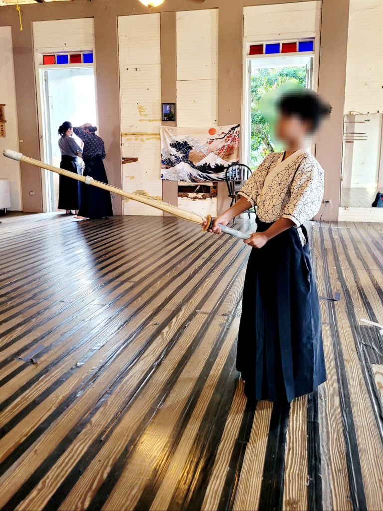 A young boy stands on a wooden floor, wearing a traditional kendo uniform with a patterned white top and dark pleated hakama pants. He holds a kendo bamboo sword in kamae stance. Behind him, a wall features open doors and a tapestry with a wave and Mount Fuji design.