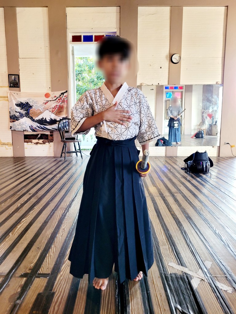 A young boy stands barefoot on a wooden floor, wearing a traditional kendo uniform with a patterned white top and dark pleated hakama pants. He holds one hand to his chest, and a kendo sword with a yellow and black handle rests in his other hand. Behind him, a wall features mirrors, a clock, and a tapestry with a wave and Mount Fuji design.